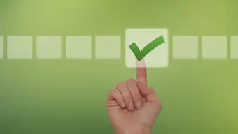 Grafika dekoracyjna. Dłoń z wyciągniętym palcem wskazującym wskazuje na zielony haczyk umieszczony w kwadracie. Tło jest zielone, a obok zaznaczonego kwadratu znajduje się kilka pustych kwadratów ułożonych poziomo.