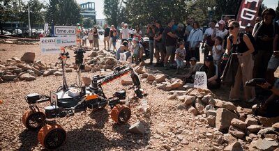 Łazik marsjański porusza się po kamienistym torze podczas zawodów European Rover Challenge. Wokół toru stoją widzowie.