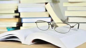 Grafika dekoracyjna: okulary położone na otwartej książce; w tle stos z książkami