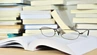 Grafika dekoracyjna: okulary położone na otwartej książce; w tle stos z książkami