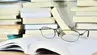 Eyeglasses laid on an open book on a table. Stacks of books in the background.