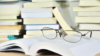 Eyeglasses laid on an open book on a table. Stacks of books in the background.