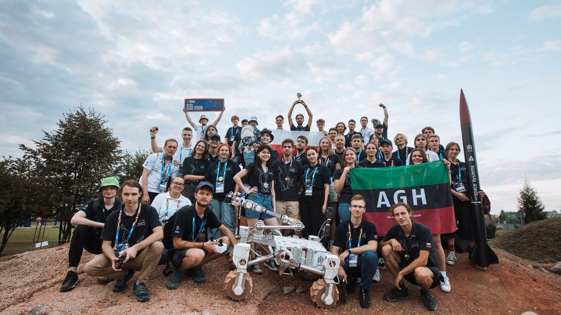 Grupa ok 30 osób - zespół AGH Space Systems, w rękach trzymają flagę AGH. Przed nimi stoi łazik Kalman, stojąca po prawej dziewczyna trzyma czarną rakietę z napisem AGH Space System