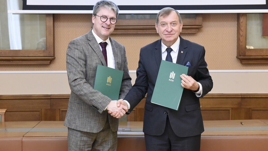 Two men dressed elegantly, shaking hands, and holding document folders with the logo of the AGH University.