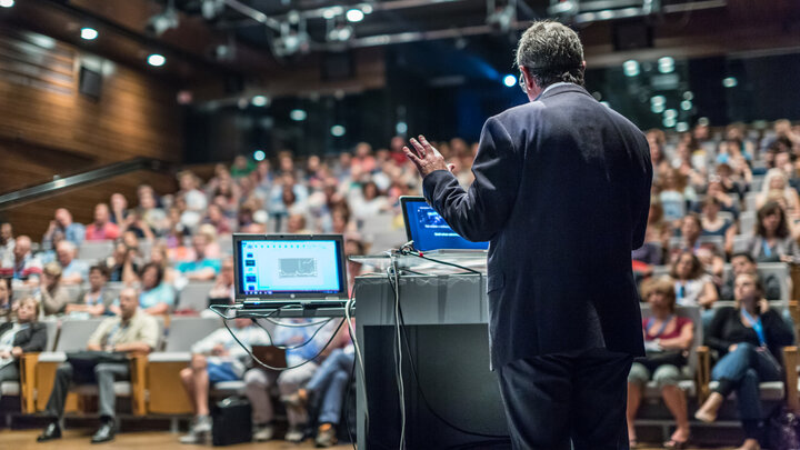 Photo from a lecture in a large auditorium. A male speaker in the centre with numerous people in the audience.