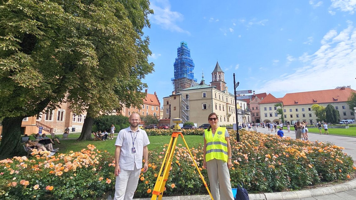 A woman and a man standing at a geodetic tripod against the Wawel buildings.