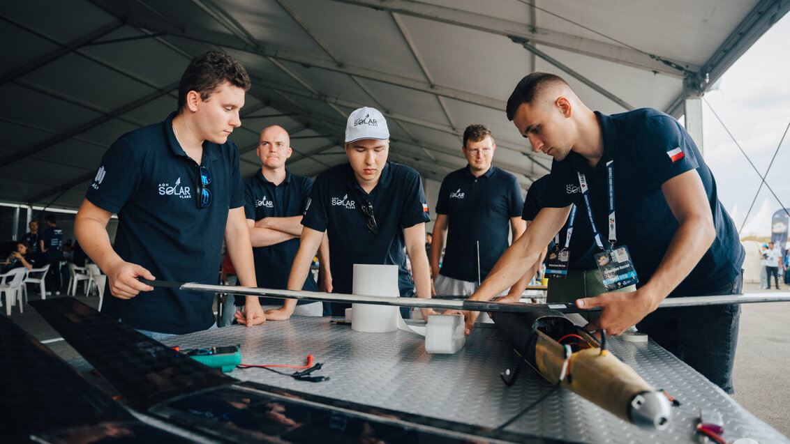 Five young men, members of the AGH Solar Plane student research club. They are gathered around a large working surface on which lies a plane model. All are wearing navy-blue polo shirts with a white inscription that says: ‘AGH Solar Plane’ and the Polish flag and the AGH UST logo.