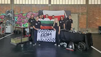 Na zdjęciu grupowe zdjęcie zespołu AGH Racing w hali zawodów. Na stołach rozłożone części konstrukcyjne bolidu. Osoby na zdjęciu trzymają w dłonicach flagi Polski i AGH.