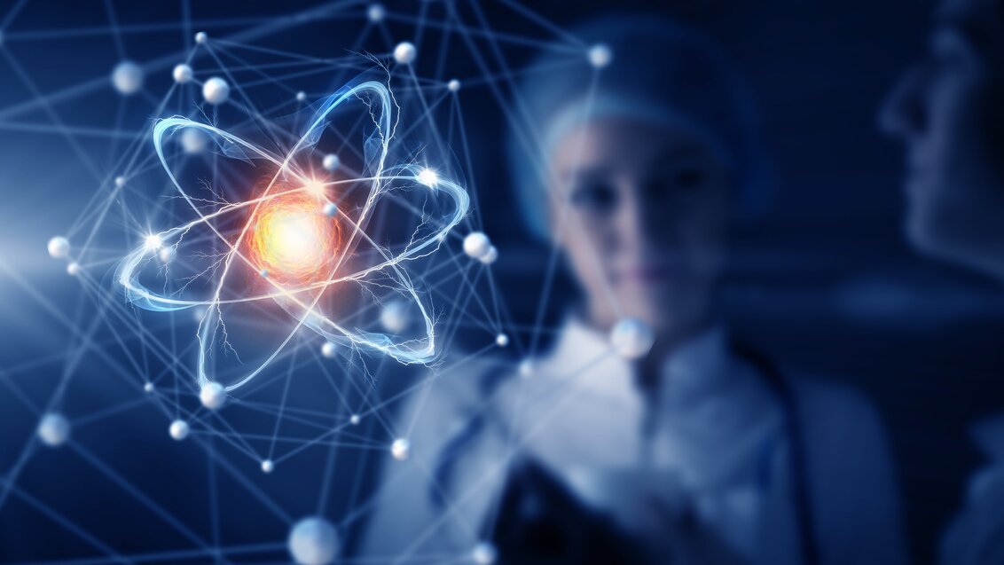 Na zdjęciu młoda kobieta-naukowiec ubrana w biały fartuch i ochronny czepek na głowie patrzy na atom widoczny na wirtualnym ekranie.