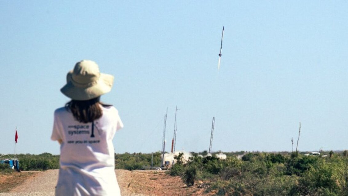 Widok wznoszącej się ku górze rakiety, tuż po starcie. Lot rakiety obserwuje osoba, stojąca tyłem do obiektywu aparatu.