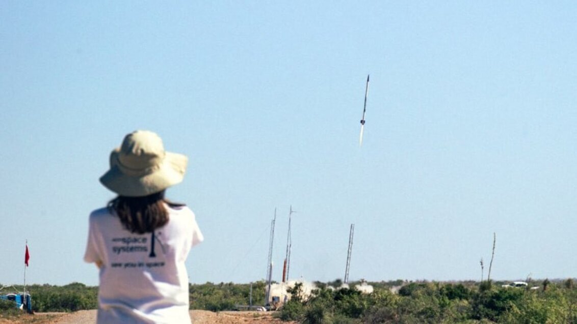 Widok wznoszącej się ku górze rakiety, tuż po starcie. Lot rakiety obserwuje osoba, stojąca tyłem do obiektywu aparatu.