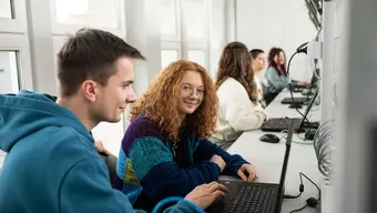 Grupa osób pracuje przy komputerach w sali wyposażonej w stanowiska komputerowe.