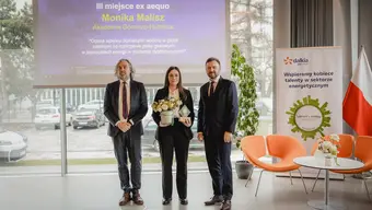 Na zdjęciu laureatka trzyma w dłoniach kwiaty i pamiątkowa statuetkę. Obok niej dwóch mężczyzn w garniturach. Za nim ekran wyświetlający planszę z tytułem pracy, imieniem i nazwiskiem autorki, nazwa uczelni oraz dopiskiem "III miejsce ex aequo".