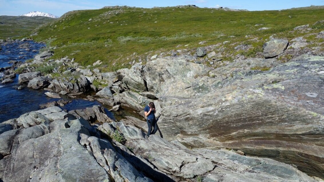 Doktorantka AGH w trakcie badań terenowych stoi na skałach przy linii brzegu. Z lewej strony widać wodę, w górnej części zdjęcia skały porośnięte są trawą.