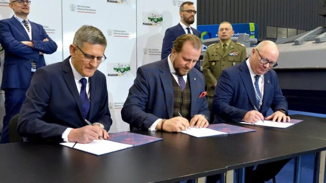 Three men in suits sitting at a table signing three copies of a document.