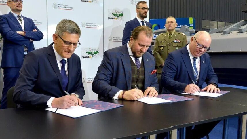 Three men in suits sitting at a table signing three copies of a document.
