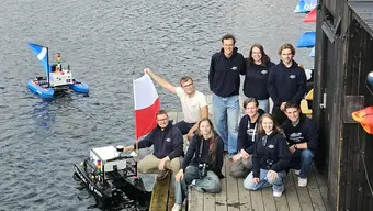 A group of young people in dark hoodies, some standing, some squatting, in a dock, nearby water. A student is holding a Polish flag. Two boats are floating on the water.