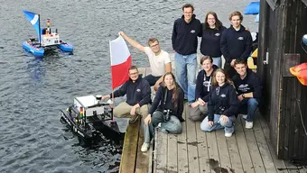 Grupowe zdjęcie (dziewięć osób) zespołu AGH Solar Boat wykonane na drewnianym pomoście nad wodą. Jedna z osób trzyma polską flagę. W tle, na wodzie unoszą się dwie małe łodzie autonomiczne.
