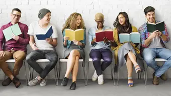  Sześciu studentów siedzi w rzędzie na krzesłach. W rękach trzymają kolorowe zeszyty, uśmiechają się. 