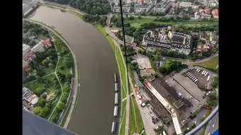 Panorama Krakowa z balonu widokowego. W centrum zdjęcia rzeka Wisła, zakręca w lewą stronę. Na lewym oraz prawym brzegu rzeki widać tereny zielone, czubki drzew oraz dachy licznych budynków.