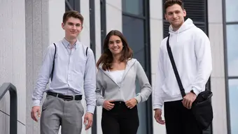 Dwóch studentów i jedna studentka (pośrodku) stoją obok siebie. Za nimi widoczny fragment elewacji jednego z budynków na kampusie.