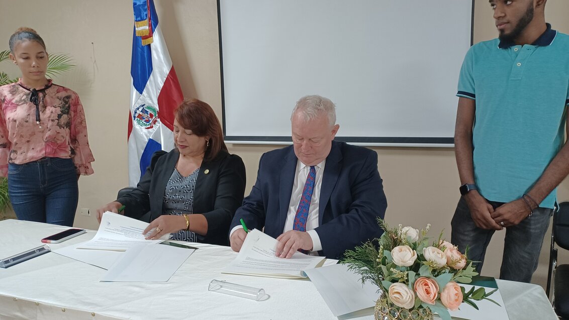 Na zdjęciu podpisanie umowy AGH – UTECO. Za stołem siedzą od lewej Rektor Marilyn Díaz i Prorektor Rafał Wiśniowski, podpisują dokumenty. Z lewej strony stołu stoi młoda kobieta, a z prawej młody mężczyzna. 