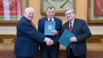 Trzech mężczyzn w garniturach stoi obok siebie, trzymając teczki z dokumentami; podają sobie rękę na znak zawarcia porozumienia.