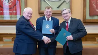An image of three men in business attire shaking hands and holding green brochures.