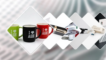 Grafika dekoracyjna przedstawiająca serię romboidalnych ramek, z których każda prezentuje inny gadżet promocyjny z logotypem „I ♥ AGH” lub „AGH”. Od lewej znajdują się: trzy kubki w kolorach zielonym, czarnym i czerwonym; długopis w etui; smycze z przypiętymi brelokami; zestaw długopisów i ołówków; oraz pendrive. Tło rozmyte, utrzymane w delikatnych kolorach.
