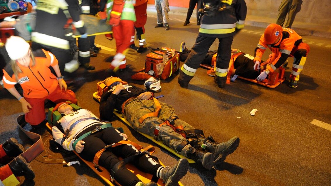 A rescue mission in a tunnel: the victims are lying on stretchers, immobilised with straps. Around them, there are medics and fire fighters. Medical bags are scattered on the road.