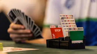 Na zdjęciu stolik do gry w brydża z przygotowaną do gry talią kart. W tle widoczna dłoń, trzymająca rozłożone karty.