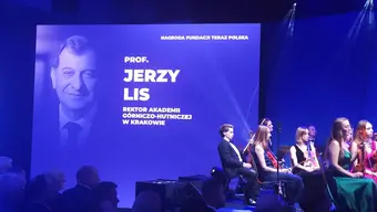 The image shows a formal event. On the left side, there is a large screen displaying a photo of a man with text: Prof. Jerzy Lis, Rector of AGH University. In front of the screen, several people are seated on stage, facing the audience.