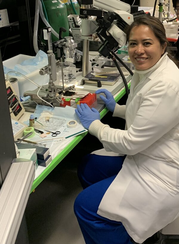 Dr Gloria Lesly Jimenez Miranda podczas pracy w laboratorium badawczym. Naukowczyni w białym fartuchu siedzi przy stanowisku wyposażonym w specjalistyczną aparaturę laboratoryjną i mikroskop.
