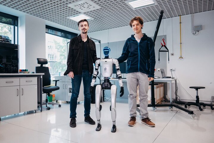 Dwóch naukowców stoi po bokach humanoidalnego robota z logo „AGH” w jasnym laboratorium wyposażonym w sprzęt i stanowiska badawcze.