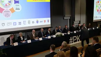 Sala konferencyjna z długim stołem, przy którym siedzi kilkanaście osób. W tle ekran z grafiką wydarzenia.