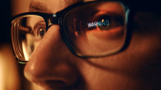 Zdjęcie zbliżenia twarzy młodego mężczyzny w okularach. W jednym ze szkieł odbija się napis "warning".