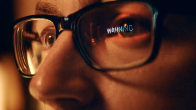 A man's face with glasses which reflect an inscription that says "WARNING".