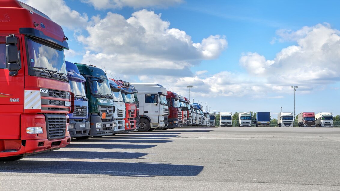 The photo shows a large parking lot. On the left and in the background of the photo, there are lorries parked in a row. The middle of the lot is empty. The sky is blue with a few white clouds.