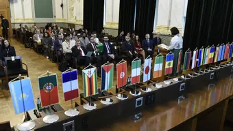 The image shows a long table displaying small flag banners of various countries, symbolising international representation. The room is the AGH University assembly hall. In the backgroung we may see its interior and the audience, the attendees of the conference. There is a woman speaking at the pulpit.