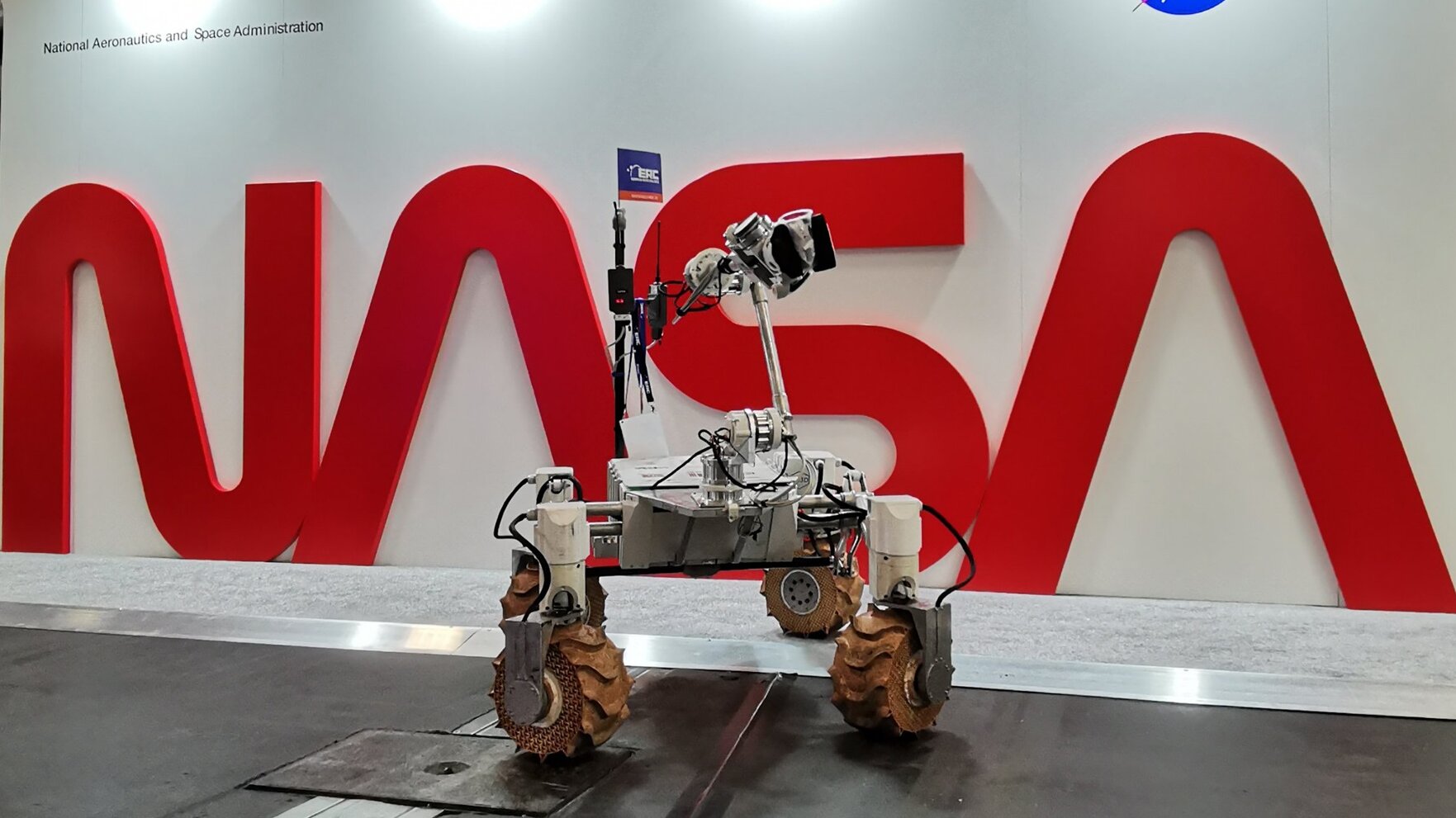 Na tle białej ściany z dużym czerwonym napisem NASA stoi robot Kalman - platforma z szeroko rozstawionymi masywnymi terenowymi kołami i osprzętowaniem badawczym. 