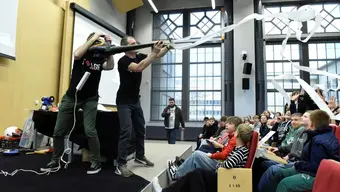 Dwóch mężczyzn na scenie trzyma dmuchawę skierowaną w stronę publiczności zgromadzonej w dużej sali. Młodzież siedzi lub stoi, łapiąc fragmenty papieru, który leci w ich stronę.