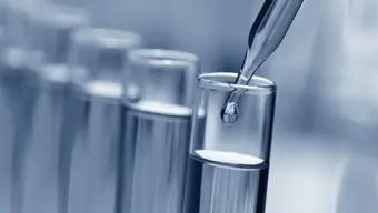 Pipeta laboratoryjna upuszczająca kroplę przezroczystej cieczy do probówki. W tle widoczne są kolejne cztery probówki z cieczą.