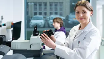 Zdjęcie. Na pierwszym planie młoda kobieta w fartuchu laboratoryjnym trzyma dłonie na obiektywie mikroskopu. Uśmiecha się. Na drugim planie widać młodego mężczyznę siedzącego przed monitorem komputera.