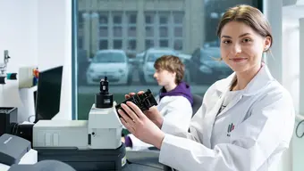 Image of a female scientist in a lab coat with the logo of the AGH University who is carefully adjusting a microscope in a well-lit laboratory. Another individual, also in a lab coat, is working at a computer in the background.