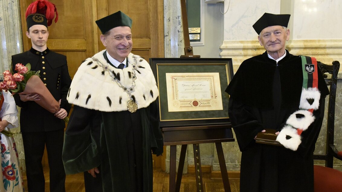 Professor Jan Kiciński and the AGH University Rector stand next to each other. Between them is a stand with the commemorative diploma. Behind the man on the right is another man in a mining uniform. He is holding a bouquet in his hands. The photo was taken during the ceremony in the AGH University Assembly Hall.