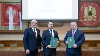 The photo shows the signers of the agreement. Three smiling men in suits stand next to each other. They are holding document folders.