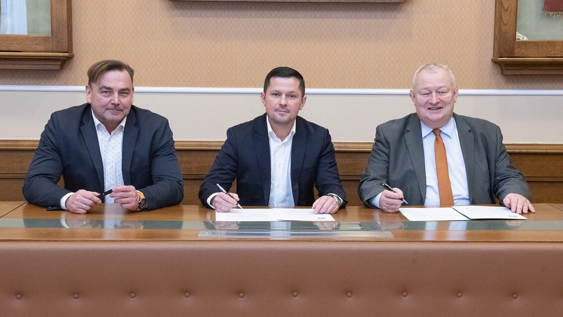 Na zdjęciu sygnatariusze umowy (trzej mężczyźni) - siedzą za stołem, przed nimi dokumenty z umową, w dłoniach trzymają długopisy. Zdjęcie wykonane w sali konferencyjnej AGH.