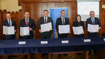 Zdjęcie grupowe upamiętniające podpisanie porozumienia. Sześć osób stoi przy stole, trzymając otwarte dokumenty.