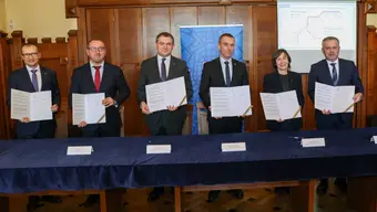 A group of five men and a woman in an official setting. Each of them is holding an opened document, smiling, standing behind a table with name cards.