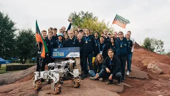 Competition grounds. The Kalman rover, behind it a group of about 30 people from the AGH Space Systems. They have medals around their necks. Two people in the first row hold a large prize voucher and someone raises the award statuette. Two people hold AGH UST-coloured flags.